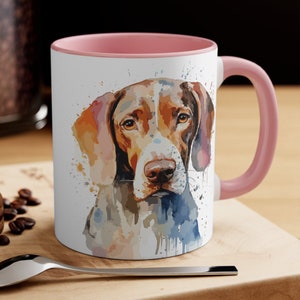 May include: A pink and white ceramic mug with a watercolor illustration of a brown and white dog. The dog's head is facing forward and its eyes are looking directly at the viewer. The background is a white watercolor wash with splashes of color.