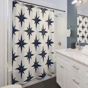 May include: A white shower curtain with a navy blue starburst pattern. The curtain hangs from a dark metal rod in a bathroom with white tile walls, a white vanity, and a dark gray floor. A mirror reflects the shower curtain.