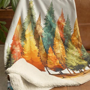 May include: A soft, white blanket with a colorful forest scene. The design features watercolor-style trees in shades of orange, yellow, green, and brown. The blanket is draped over a brown leather couch, showcasing its cozy sherpa backing.