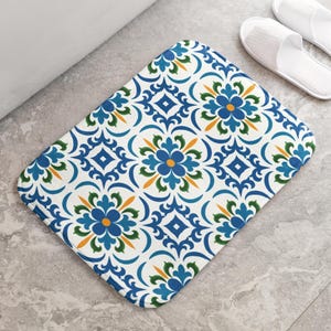May include: Rectangular bath mat with a white background and a repeating pattern of blue and green floral and geometric designs. The mat is placed on a gray tiled floor, with white slippers visible in the background.