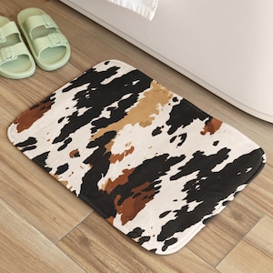 Puede incluir: Una alfombra de baño con estampado de piel de vaca en marrón, negro y blanco. La alfombra está hecha de un material suave y absorbente, ideal para añadir un toque de estilo rústico a tu baño.