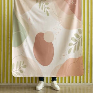 Puede incluir: Una manta suave con un diseño abstracto en tonos rosa, melocotón y verde. La manta presenta formas orgánicas, ramitas en forma de hojas y acentos en forma de estrella sobre un fondo blanco. La manta está sostenida por una persona que lleva zapatillas blancas.