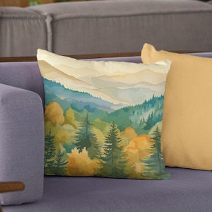May include: A throw pillow with a watercolor painting of a mountain range with a forest in the foreground. The mountains are a light blue and the forest is a mix of green and yellow.