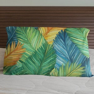 May include: A rectangular pillow with a vibrant watercolor design of tropical palm leaves. The leaves are in shades of green, blue, yellow, and orange. The pillow is placed on a bed with a light-colored comforter and a brown headboard.