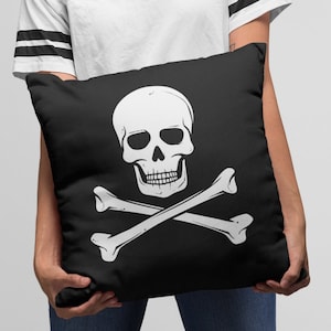 May include: A black pillow with a white skull and crossbones design. The pillow is being held by a person wearing a white t-shirt with black stripes and blue jeans.