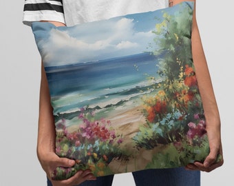 May include: A cushion displaying a coastal landscape painting. The artwork shows a beach scene with blue sea, a sandy shore, and colourful flowers. The cushion is held by a person wearing a white t-shirt and blue jeans.