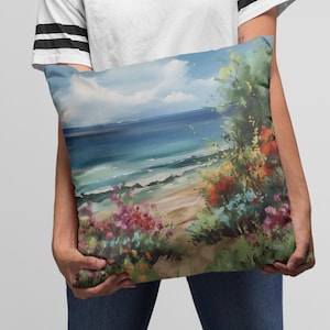 May include: A decorative pillow featuring a coastal landscape painting. The artwork depicts a beach scene with blue ocean water, a sandy shore, and colorful flowers. The pillow is held by a person wearing a white t-shirt and blue jeans.