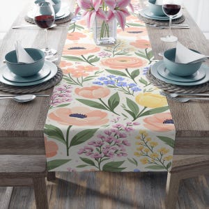 Può includere: Una tovaglia floreale con sfondo crema e un motivo di fiori color pesca, gialli e viola. Foglie e steli verdi sono sparsi nel design. La tovaglia è posta su un tavolo di legno con delle posate.