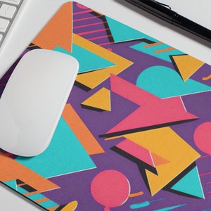 May include: A colorful geometric mouse pad with a purple background, featuring a variety of shapes in shades of orange, yellow, pink, blue, and turquoise.