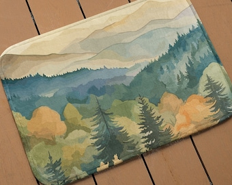 Puede incluir: Una pintura de acuarela de un paisaje de montaña con un bosque en primer plano. Las montañas están en el fondo y son de color azul claro. El bosque es una mezcla de árboles verdes y marrones. La pintura está sobre un fondo beige.