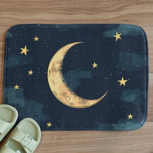 Può includere: Un tappetino da bagno blu con una luna crescente e stelle dorate. La luna è al centro del tappetino e le stelle sono sparse intorno.