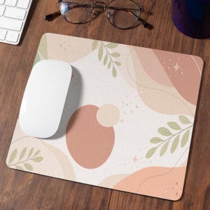 Könnte beinhalten: Rechteckiges Mauspad mit cremefarbenem Hintergrund und abstrakten Formen in Pfirsich, Beige und Hellgrün. Eine weiße Computermaus liegt auf dem Pad. Das Design umfasst Blatt- und Sternakzente.