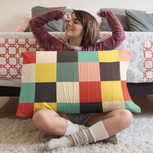 May include: A rectangular pillow with a colorful patchwork design. The pillow features a grid of squares in various colors, including red, yellow, teal, and brown. The pillow is resting on a person's lap, who is sitting on a white rug.