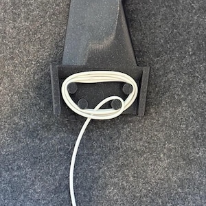 May include: A black phone charging stand with a white charging cable wrapped around it. The stand has a circular, silver-rimmed charging pad on top.