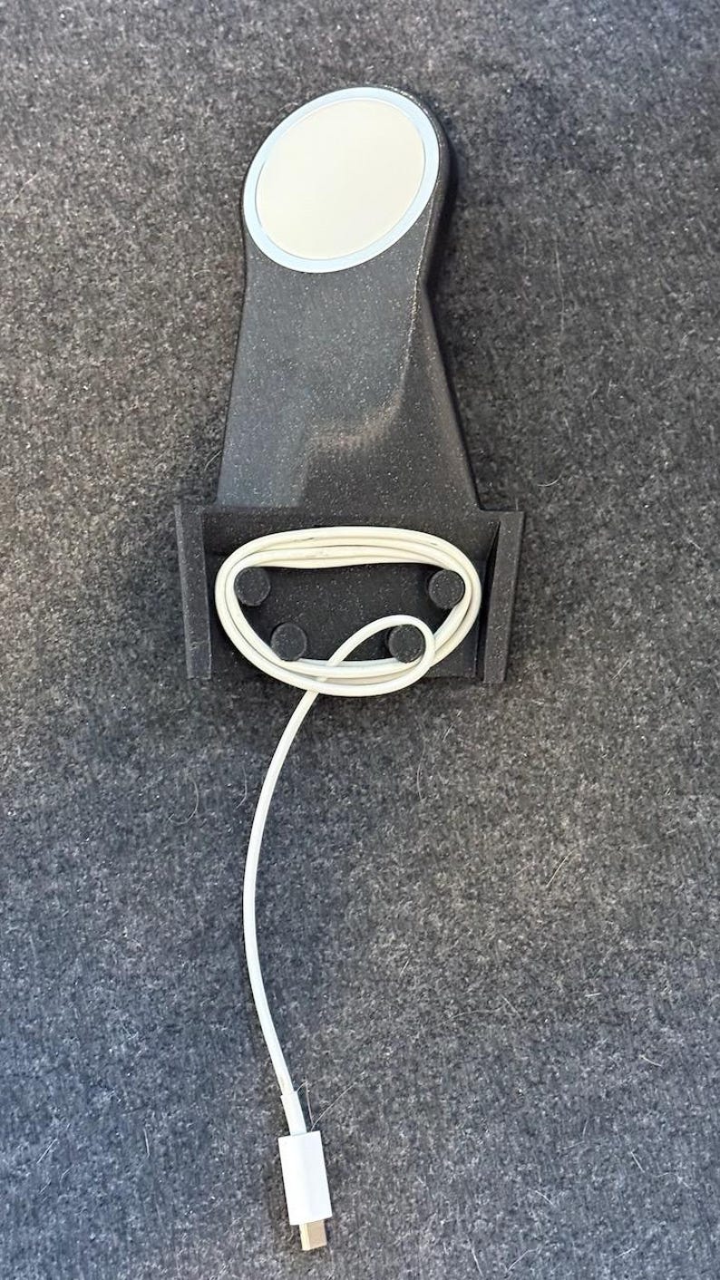 May include: A black phone charging stand with a white charging cable wrapped around it. The stand has a circular, silver-rimmed charging pad on top.