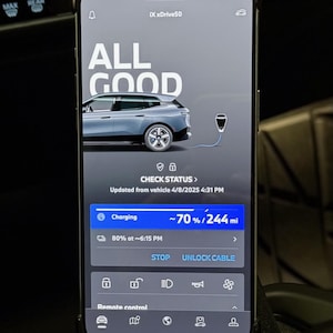 May include: A smartphone screen displaying a car charging status. The screen shows "ALL GOOD" with a picture of a car charging. The car is a silver BMW iX xDrive50. The screen shows the car is charging at 70% and has 244 miles of range. The screen also shows the time the car will be fully charged, 6:15 PM. The screen has buttons for "STOP" and "UNLOCK CABLE".