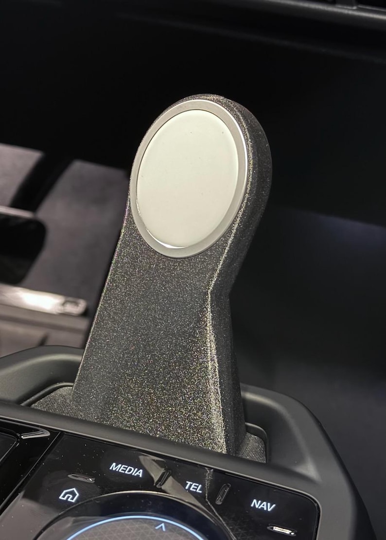 May include: A black gear shifter with a silver ring around a white button. The shifter has the words "MEDIA", "TEL", and "NAV" on the bottom.