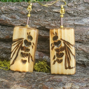 May include: A pair of wooden earrings with a burnt wood design of a bee. The earrings are on a wooden surface with green moss.