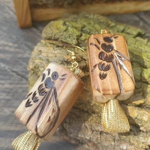 May include: A pair of wooden earrings with a bee design and gold tassels. The earrings are made of natural wood and have a rustic look.