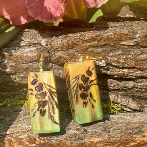 May include: A pair of handmade earrings with a floral design. The rectangular earrings are gold with a green ombre effect at the bottom. Black floral designs are etched on the front. The earrings have gold hooks and small turquoise beads.