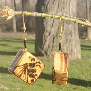 May include: A pair of gold and black dangle earrings with a square-shaped ceramic charm. One charm features a black and white design of a bee, while the other charm is plain.