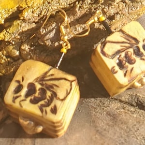 May include: A pair of square-shaped earrings with a carved design of a bee on each earring. The earrings are made of a light brown material and have gold-colored hooks.