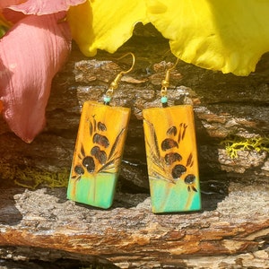 May include: A pair of dangle earrings with a rectangular shape. The earrings are made of wood and have a yellow, orange, and green color scheme. The earrings feature a floral design with black accents.