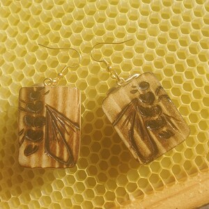 May include: A pair of gold earrings with a rectangular wooden charm featuring a detailed image of a bee on a honeycomb background.