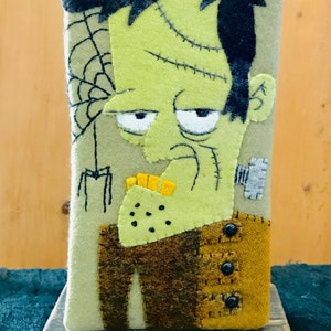 May include: A handmade Halloween-themed felt decoration featuring a stylized Frankenstein's monster. The monster has a green face, black hair, and stitched details. The design includes a spider web and is mounted on a wooden base.