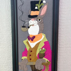 May include: A felt wall hanging of a rabbit wearing a top hat, bow tie, and a pink jacket. The rabbit is holding a pipe and has a frog in its pocket.