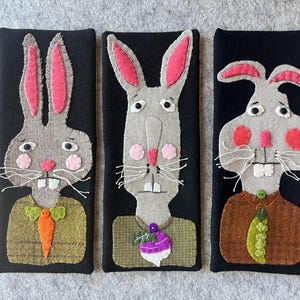 May include: Three felt bunny portraits on black backgrounds. Each bunny has gray fur, pink inner ears, and a unique outfit. One wears a green bow tie with a carrot, another a purple turnip, and the last a green pea pod.