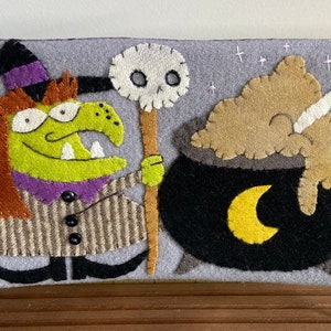 May include: A gray felt Halloween decoration featuring a green witch with orange hair, a black hat, and a striped coat. A black cauldron with a yellow crescent moon and a bone are also depicted. The witch holds a staff with a skull.