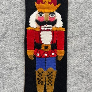 May include: A decorative, rectangular Christmas ornament featuring a colorful nutcracker design. The nutcracker is dressed in a red jacket, blue pants, and gold boots, with a white beard and a gold crown. The background is black.