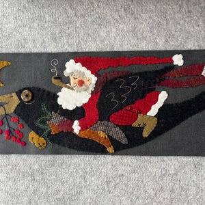 May include: A whimsical felt art piece depicting Santa Claus riding a black crow across a night sky. Santa wears a red suit and smokes a pipe. The crow carries a sprig of berries and a crescent moon is in the background.
