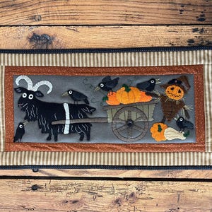 May include: A rectangular Halloween-themed fabric wall hanging. It features a black goat pulling a cart filled with pumpkins, with crows perched on the goat and cart. A scarecrow is also present. The border has striped and dotted patterns.