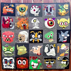 May include: A handmade quilt featuring a grid of colorful monster faces. Each square showcases a unique, appliqued monster design with varying colors, shapes, and expressions. The quilt is bordered with a black fabric.