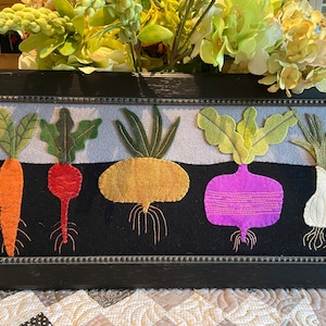 May include: A framed needlepoint artwork depicting five different vegetables: a carrot, a beet, a turnip, a rutabaga, and an onion. The vegetables are all embroidered in different colors and textures, and they are arranged in a row against a black background.
