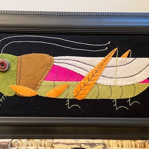 May include: A framed felt artwork depicting a green grasshopper with orange legs and wings. The grasshopper has a brown and pink body and a large red button for an eye.