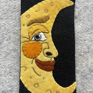 May include: A black rectangular felt case featuring a smiling yellow crescent moon with a face. The moon has a brown eyebrow, an orange cheek, and a dark brown mouth. The moon's face is detailed with a white eye and small dots.