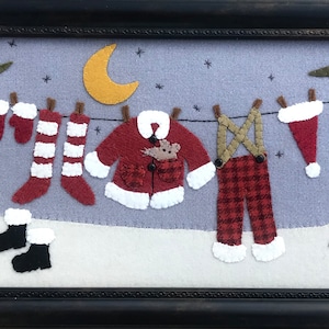 May include: A felt Christmas scene with a clothesline hanging Santa's clothes, including a red and white striped stocking, a red jacket with a white trim, red and black plaid pants, and a red hat. The scene is set against a blue sky with a yellow crescent moon and stars. A sign reads "North Pole" in brown letters.