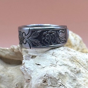 Puede incluir: Un anillo de plata con un diseño de rosa grabado. El anillo está sobre un trozo de madera a la deriva.