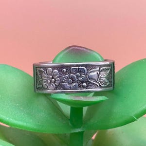 May include: A silver ring with a floral design. The ring is set against a green background.