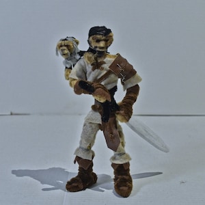May include: A brown and white felt figure of a person wearing a brown leather belt and holding a sword. The figure is wearing a white tunic and has a brown felt animal on its shoulder.