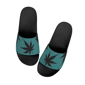 Op de afbeelding: Zwarte badslippers met een turquoise band met een herhalend zwart cannabisbladpatroon. De sandalen hebben een effen zwarte zool en een voorgevormd voetbed. Het ontwerp is eenvoudig en modern, geschikt voor casual wear.