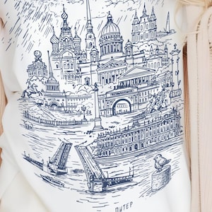 May include: White t-shirt featuring a detailed blue line drawing of a cityscape, including buildings, bridges, and a river. The design includes the word "ПИТЕР" at the bottom. The shirt has a V-neck.