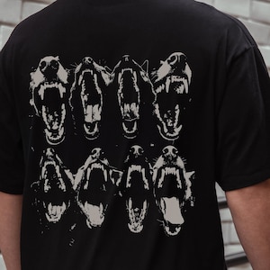 Puede incluir: Camiseta negra con un estampado gráfico de seis cabezas de perro con la boca abierta, mostrando los dientes. Las cabezas de perro están dispuestas en dos filas de tres.