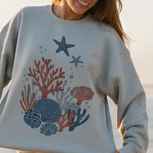 May include: A light gray sweatshirt featuring a colorful coral reef design. The graphic includes red, blue, and gray coral, two blue starfish, and bubbles. The sweatshirt has a crew neck and long sleeves.