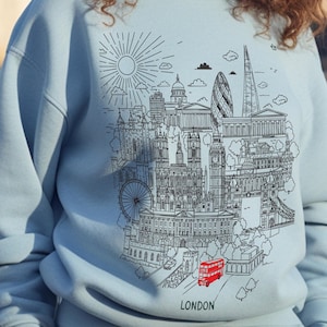 May include: Pale blue sweatshirt with a black and white illustration of London's skyline. Iconic buildings like Big Ben, the Shard, and the Gherkin are shown, along with a red double-decker bus. The word "LONDON" is printed at the bottom.