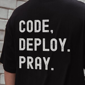 Könnte beinhalten: Schwarzes T-Shirt mit dem weißen Text "CODE, DEPLOY, PRAY." Der Text ist in einer fetten, serifenlosen Schriftart gehalten. Das T-Shirt besteht aus einem weichen Material.