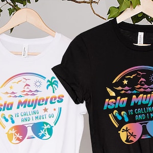 以下が含まれることがあります： 白と黒の2枚のTシャツ。それぞれにカラフルなグラフィックが施されています。「Isla Mujeres IS CALLING AND I MUST GO」の文字が虹色のアーチ状に配置され、サングラス、ヤシの木、海の要素が描かれています。